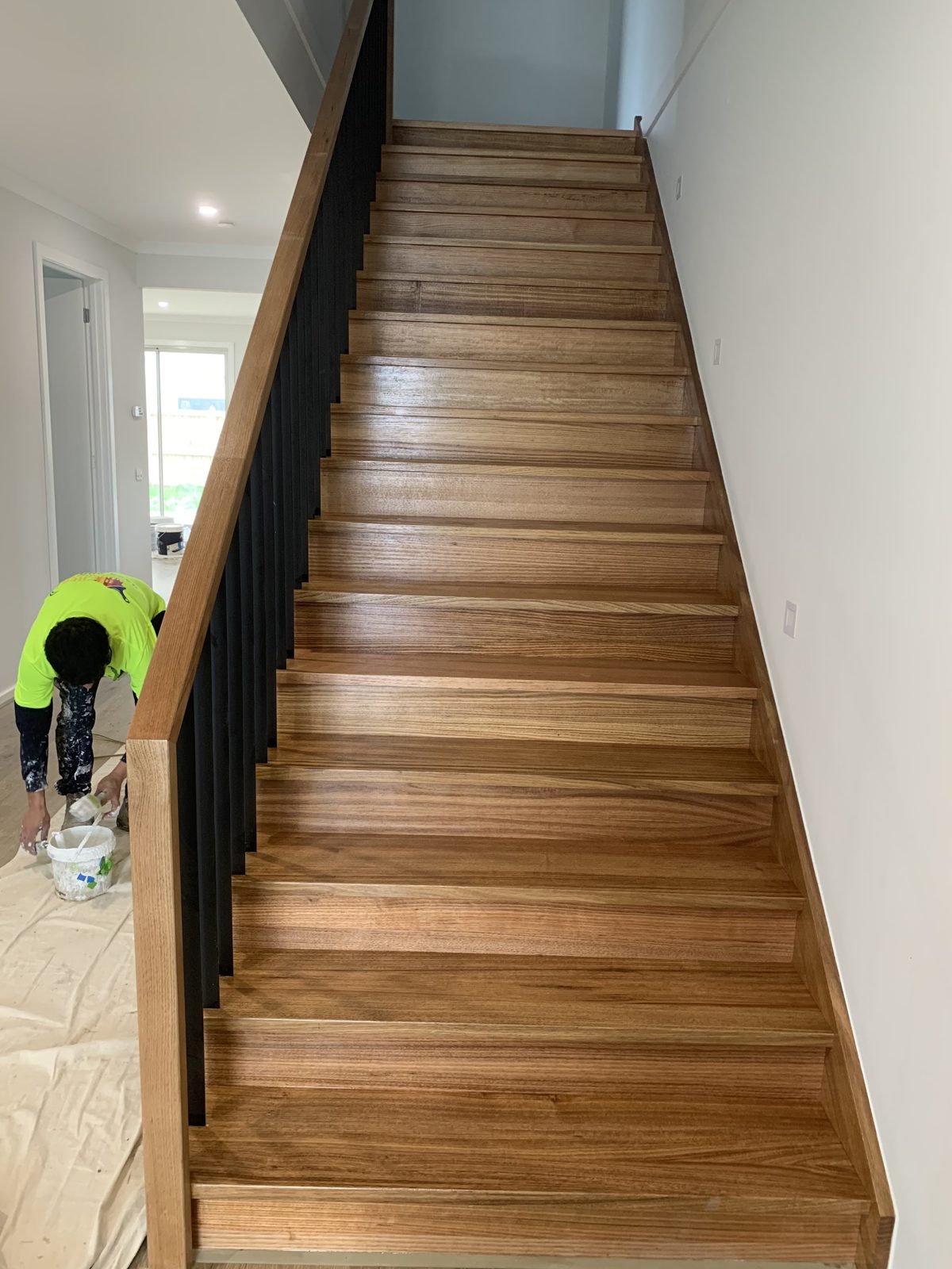 Painter in hi-vis painting a black stair balustrade beside new timber treads, floor covered with drop sheets